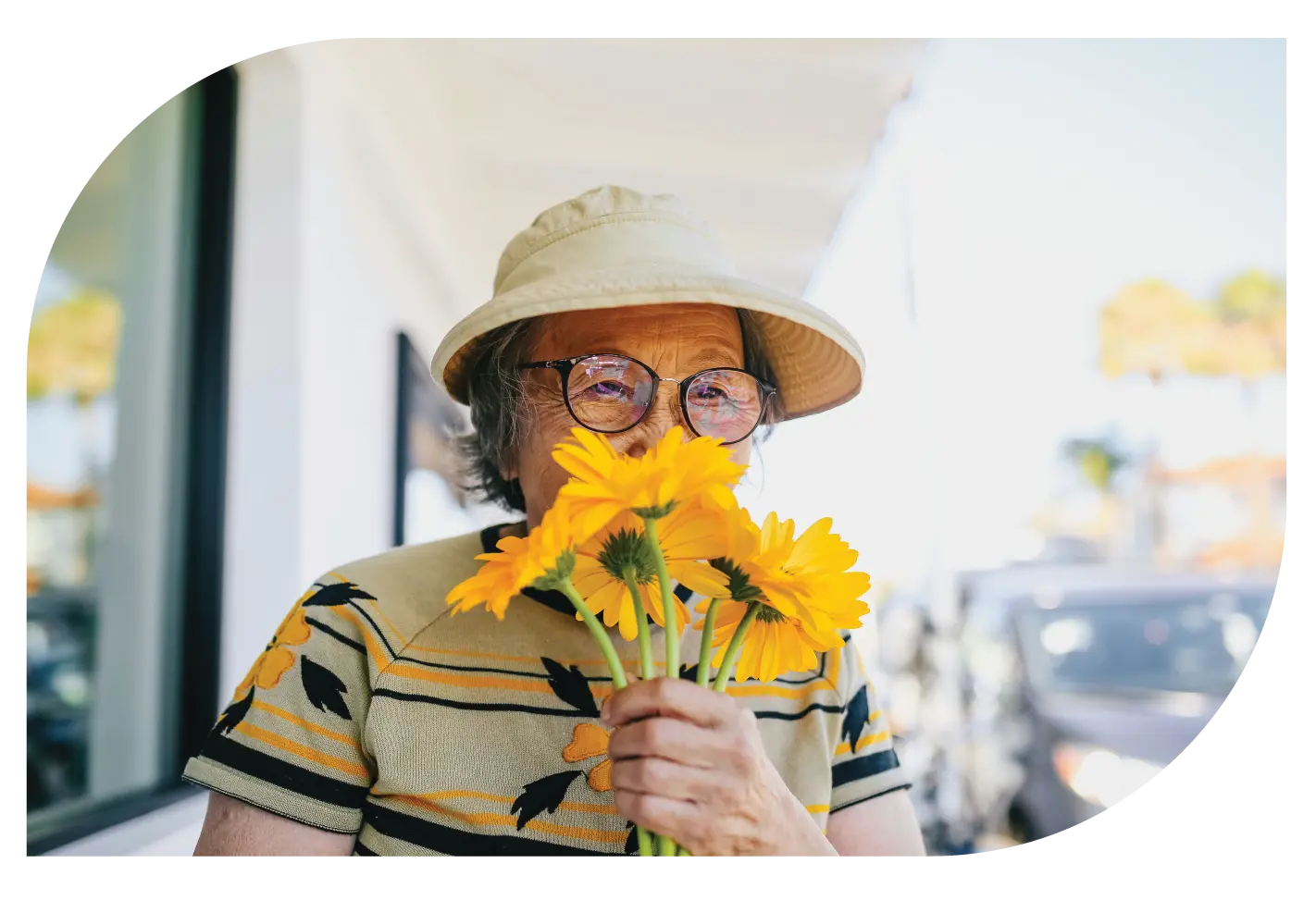 An elderly woman wearing a hat and glasses, holding a bouquet of yellow flowers.